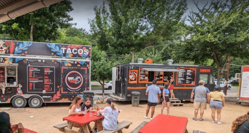 Two food trucks in a park setting, one serving tacos and the other offering various dishes, with people dining at picnic tables.