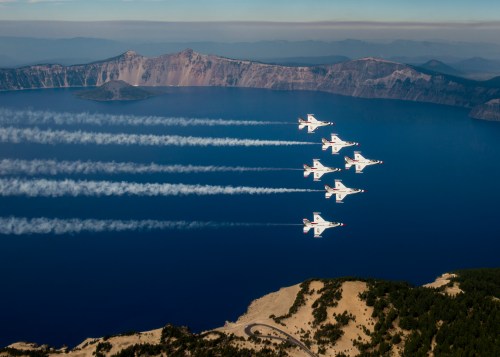 Six jets flying in formation over a deep blue lake, with a mountainous landscape in the background.