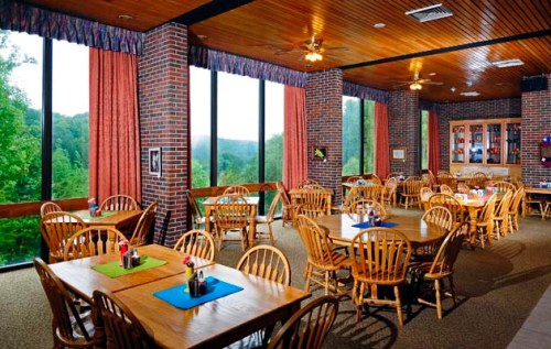 Cozy dining area with wooden tables and chairs, large windows showcasing a green landscape, and warm lighting.