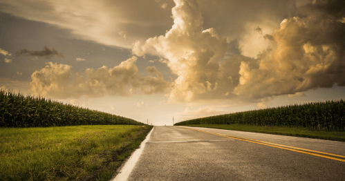 A winding road stretches through lush green fields under a dramatic sky filled with clouds.
