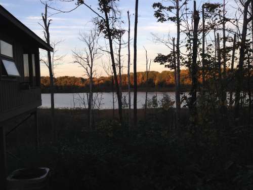 A serene view of a lake surrounded by trees at sunset, with a cabin partially visible in the foreground.