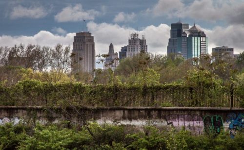 City skyline partially obscured by greenery and a graffiti-covered wall, under a cloudy sky.