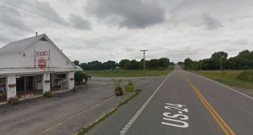 A rural road with a white building on the left, labeled "Peaches," and a grassy area on the right under a cloudy sky.