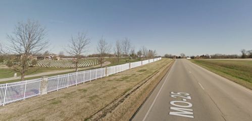 A rural road lined with a white fence, with a cemetery visible in the background under a clear blue sky.