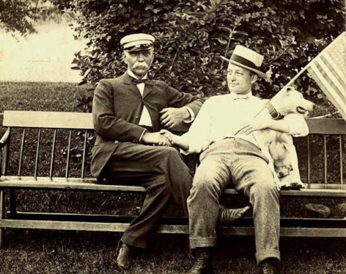 Two men in vintage clothing sit on a bench, one in a naval uniform, the other holding a dog and an American flag.