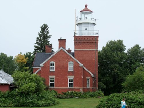 A red brick lighthouse with a white lantern room, surrounded by greenery and trees, on a cloudy day.