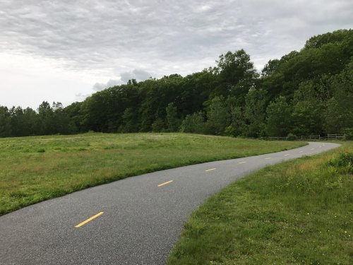 A winding path through a grassy field, bordered by trees under a cloudy sky.