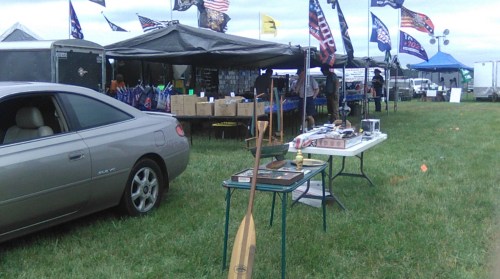 A car parked near a vendor area with tables displaying various items and flags in the background.