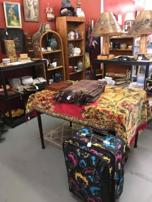 A vintage shop interior featuring a table with a colorful tablecloth, lamps, and a patterned suitcase on the floor.