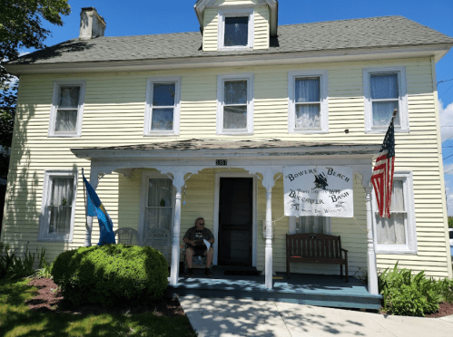 A yellow house with a porch, featuring a sign for "Bowers Beach" and an American flag, with a person sitting on the bench.