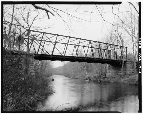 A black and white photo of a historic iron bridge over a calm river, surrounded by bare trees and rocky banks.