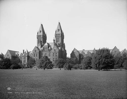 Historic Buffalo State Hospital building with twin towers, surrounded by trees and a grassy area. Black and white photo.
