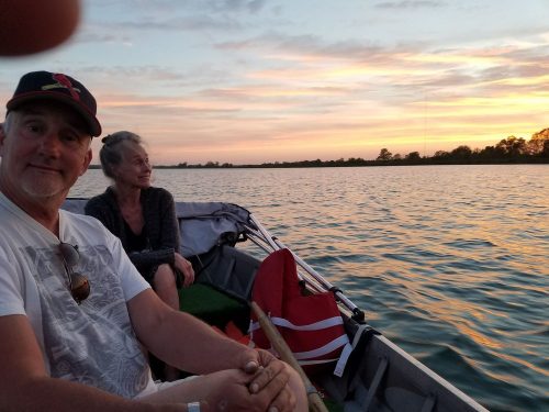 A man and woman sit in a boat, enjoying a sunset over calm waters. The scene is peaceful and serene.