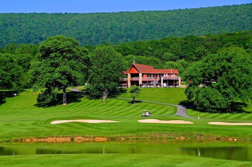 A scenic golf course with a clubhouse, lush green hills, and a calm pond in the foreground.