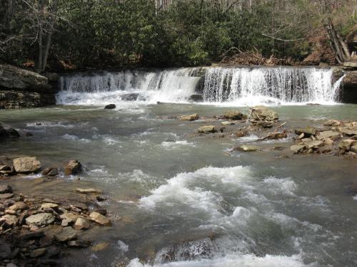A serene river flows over a small waterfall, surrounded by rocks and lush greenery in a tranquil natural setting.