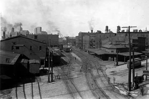 Black and white photo of a city street with buildings, smoke, and streetcar tracks, depicting an industrial area.