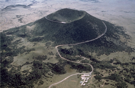 Aerial view of a volcanic cone surrounded by green landscape and a winding road leading to the summit.