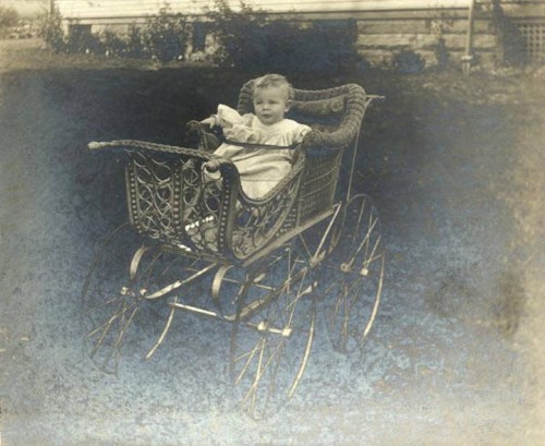 A vintage photo of a baby sitting in an ornate wicker stroller outdoors.