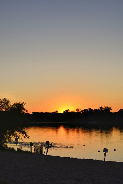 Sunset over a calm lake, with silhouettes of trees and a sandy shore in the foreground.
