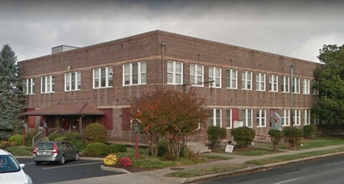 A brick building with multiple windows, surrounded by greenery and flowers, on a cloudy day.