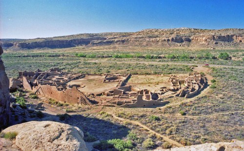 Aerial view of ancient ruins in a desert landscape, surrounded by greenery and rocky cliffs under a clear blue sky.
