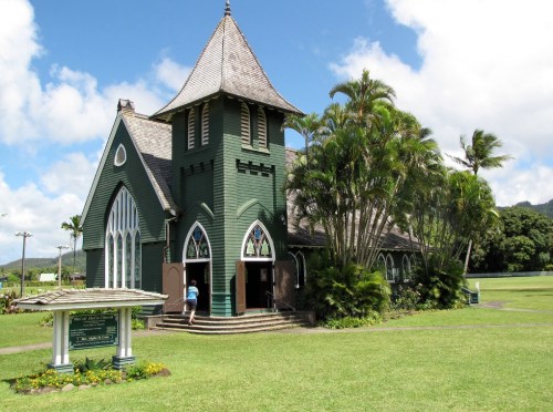A green church with a tall steeple surrounded by palm trees and a grassy area under a blue sky.