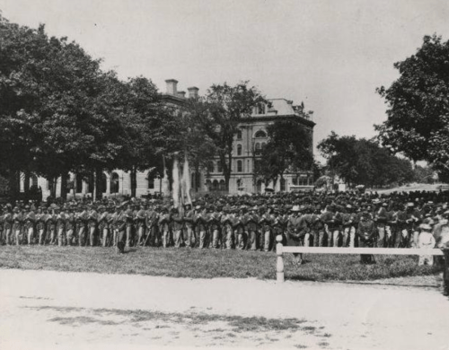 A large group of soldiers stands in formation outdoors, with trees and a historic building in the background.