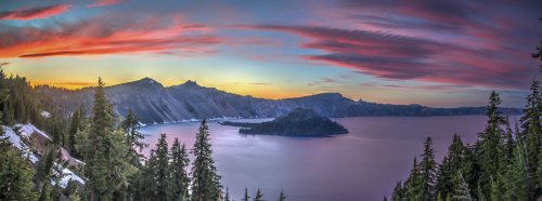 A panoramic view of Crater Lake at sunset, with vibrant colors in the sky and lush trees framing the scene.