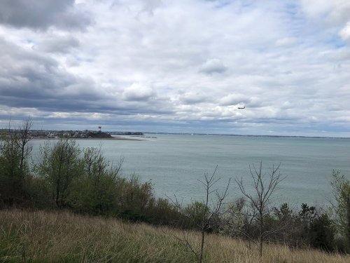 A scenic view of a calm lake under a cloudy sky, with distant land and an airplane flying overhead.