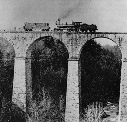 A historic black-and-white photo of a steam locomotive crossing a stone viaduct with arches.