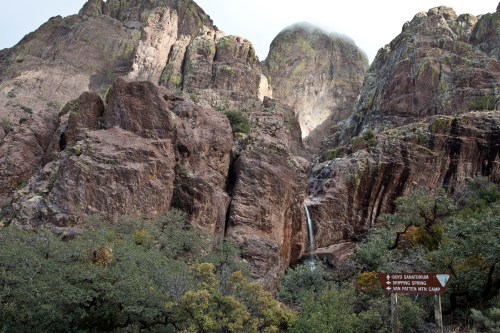 A rocky landscape with a waterfall and lush greenery, surrounded by towering mountains and a directional sign.