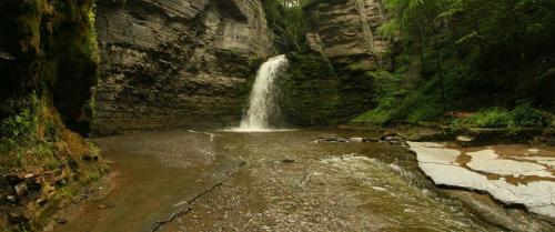 A serene waterfall cascades into a rocky pool, surrounded by lush greenery and steep stone cliffs.