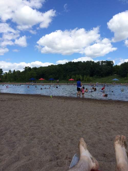 A sandy beach with people swimming in a lake, colorful umbrellas, and a blue sky with fluffy clouds.