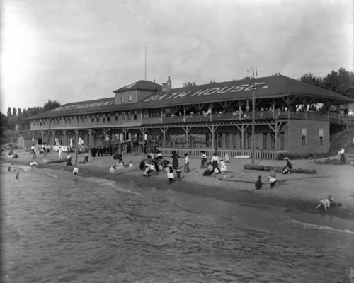 Historic black-and-white photo of a bathhouse by the beach, with people enjoying the water and relaxing on the shore.