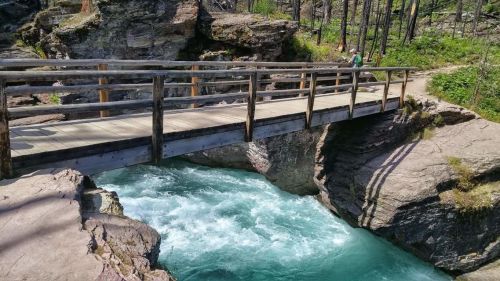 A wooden bridge spans a rushing turquoise river, surrounded by rocky terrain and greenery.