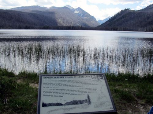 A serene lake surrounded by mountains, with a historical sign about a bomber crash in the foreground.