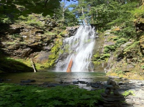 A serene waterfall cascades down rocky cliffs, surrounded by lush greenery and a calm pool, with stacked stones in the foreground.