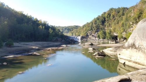 A serene river scene with a waterfall in the background, surrounded by lush green hills and rocky formations.