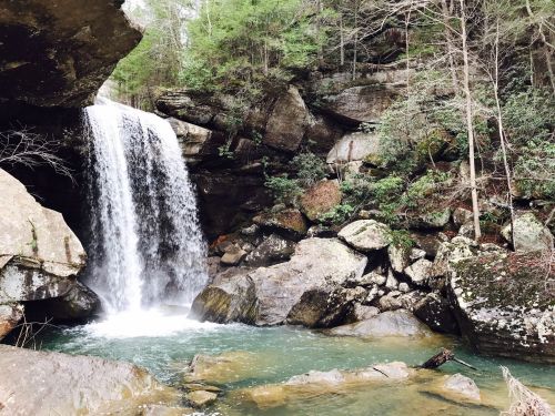 A serene waterfall cascades over rocks into a clear pool, surrounded by lush greenery and rugged terrain.