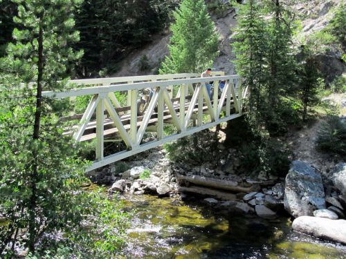 A white wooden bridge spans a clear stream, surrounded by lush green trees and rocky terrain.