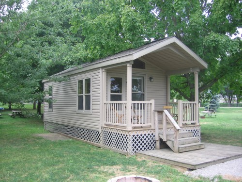 A small, beige mobile home with a porch, surrounded by green trees and grass in a park setting.