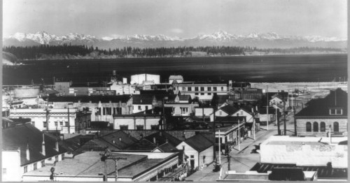 A historic black-and-white view of a town by the water, with mountains in the background and buildings in the foreground.