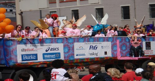 A colorful parade float with people in pink shirts, balloons, and whimsical costumes celebrating an event.
