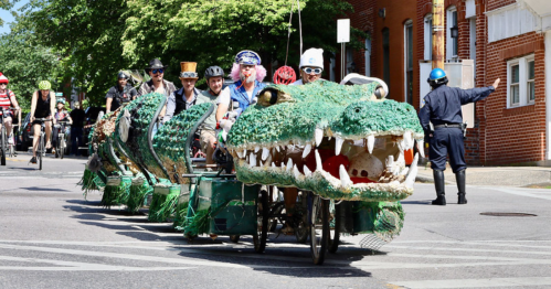 A group of people on a large, decorated bicycle resembling a crocodile, parading down a street.