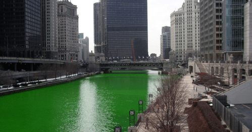 A city river dyed bright green, surrounded by tall buildings and a cloudy sky.