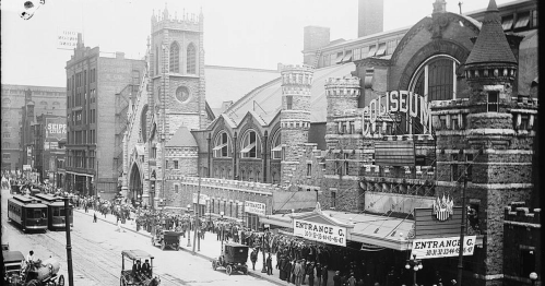 Historic black-and-white photo of a busy street with a large building labeled "Coliseum" and crowds outside.