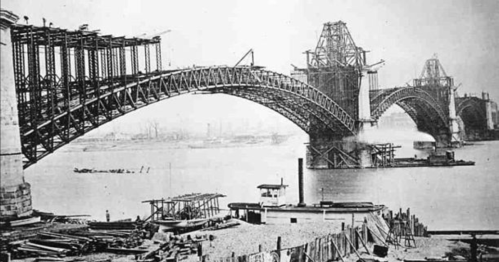 Historic black-and-white photo of a bridge under construction over a river, with scaffolding and boats nearby.