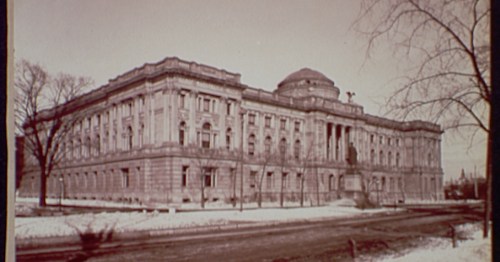Historic building with ornate architecture, surrounded by trees and snow, featuring a statue in front.