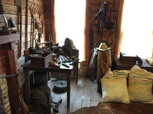 A rustic room filled with tools, wooden furniture, and various items, illuminated by natural light from two windows.