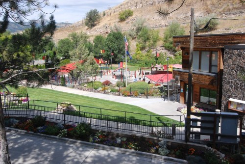 A scenic view of a park with playground equipment, gardens, and a building, surrounded by trees and hills.